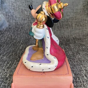 May include: A ceramic figurine of Mickey Mouse dressed as a king. He is wearing a red and gold crown, a red and white robe, and holding a gold scepter. He is standing on a purple and white platform with a brown base.