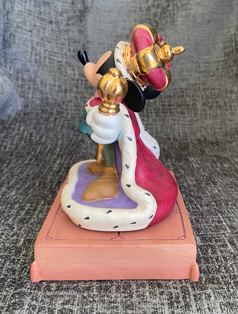 May include: A ceramic figurine of Mickey Mouse dressed as a king. He is wearing a red and gold crown, a red and white robe, and holding a gold scepter. He is standing on a purple and white platform with a brown base.