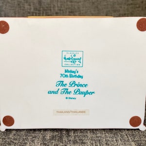 May include: White ceramic figurine base with brown felt pads on the bottom. The base has a blue and white label with the text "CLASSICS COLLECTION Mickey's 70th Birthday The Prince and The Pauper &copy; Disney THAILAND/THAILANDE".