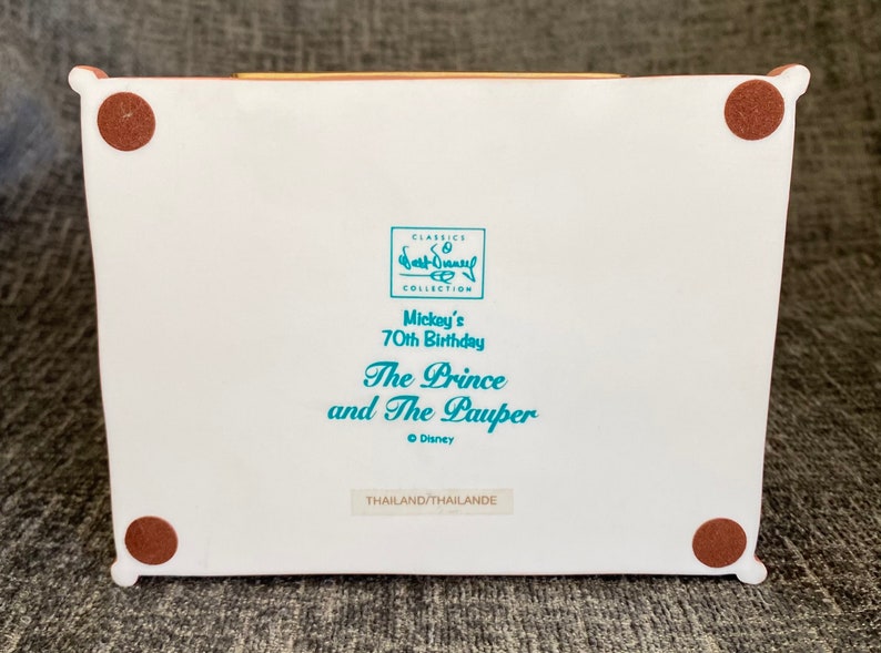 May include: White ceramic figurine base with brown felt pads on the bottom. The base has a blue and white label with the text "CLASSICS COLLECTION Mickey's 70th Birthday The Prince and The Pauper &copy; Disney THAILAND/THAILANDE".