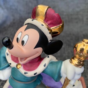 May include: A porcelain figurine of Mickey Mouse dressed as a king. He is wearing a gold crown with a red velvet lining, a blue and green robe with a gold trim, and a white ermine collar. He is holding a gold scepter.