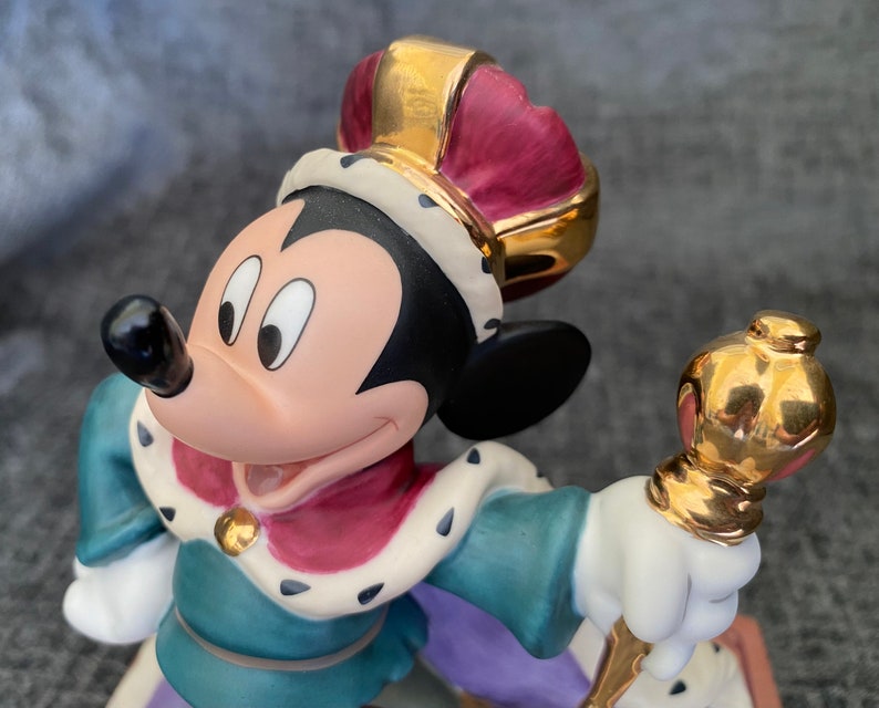 May include: A porcelain figurine of Mickey Mouse dressed as a king. He is wearing a gold crown with a red velvet lining, a blue and green robe with a gold trim, and a white ermine collar. He is holding a gold scepter.