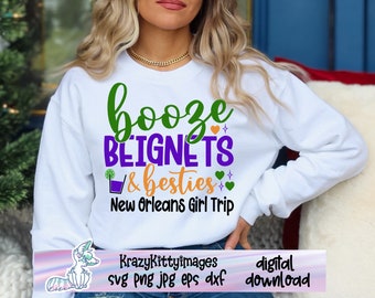Op de afbeelding: Wit sweatshirt met de tekst "booze BEIGNETS & besties New Orleans Girl Trip" in groene, paarse en oranje letters. De tekst is omringd door kleine hartjes en sterren.