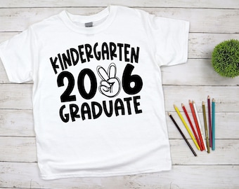 May include: White t-shirt with the text "Kindergarten 2026 Graduate" in black. The number 6 is styled with a peace sign. Colored pencils and a small green plant are on a light wood background.