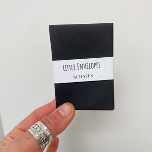 May include: A small, black package of "Little Envelopes" with the word "Scraps" underneath, held by a hand wearing multiple silver rings. The text is printed on a white label wrapped around the package.