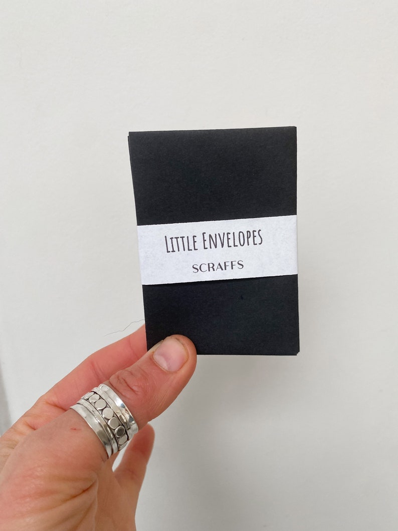 May include: A small, black package of "Little Envelopes" with the word "Scraps" underneath, held by a hand wearing multiple silver rings. The text is printed on a white label wrapped around the package.
