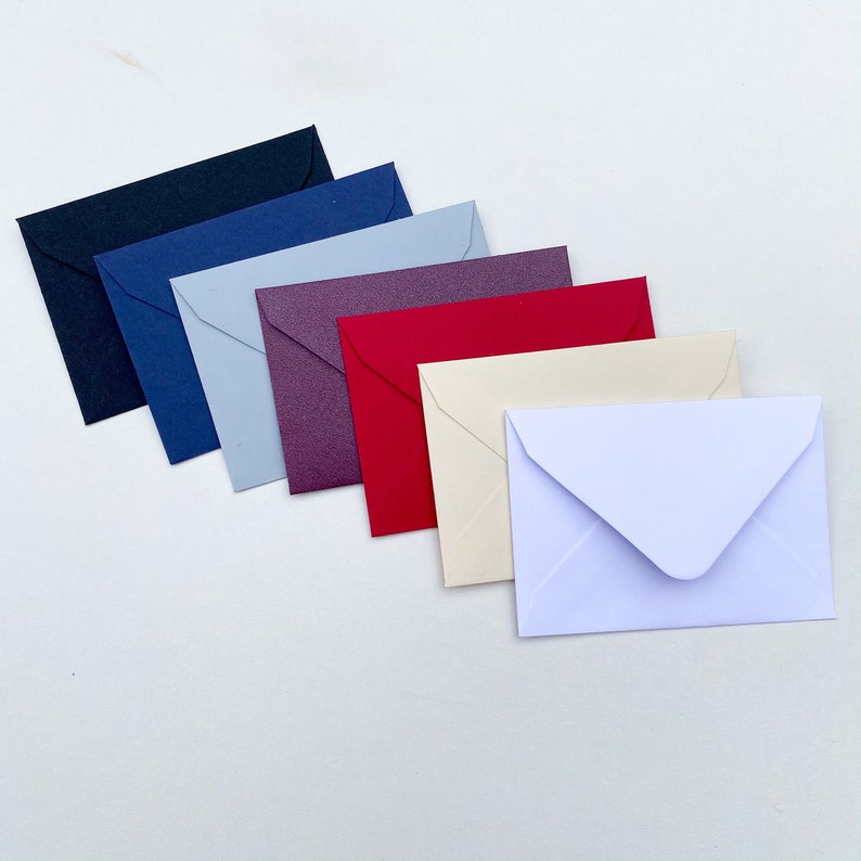 May include: A collection of small, colorful envelopes in various shades, including black, navy, light blue, purple, red, cream, and white. The envelopes are arranged in a diagonal line, showcasing their different colors and textures. Ideal for stationery and crafting.