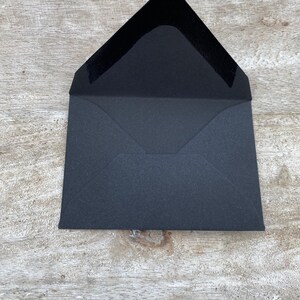 May include: An open, black envelope with a textured interior. The envelope is rectangular with a triangular flap. The interior of the flap is a smooth, dark material. The envelope is on a light-colored, wooden surface.