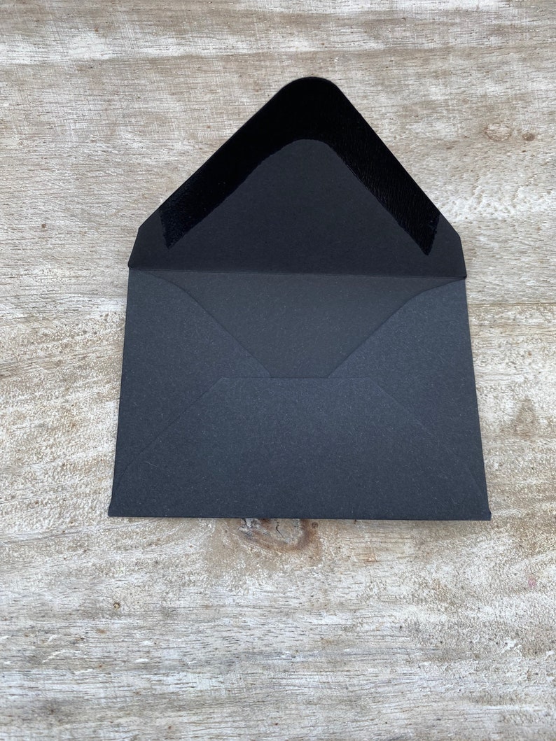 May include: An open, black envelope with a textured interior. The envelope is rectangular with a triangular flap. The interior of the flap is a smooth, dark material. The envelope is on a light-colored, wooden surface.