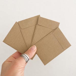 May include: A set of four small, rectangular, light brown kraft paper envelopes arranged in a fan shape. Each envelope features a pointed flap on the top edge and a smooth, matte texture consistent with standard recycled paper stationery.