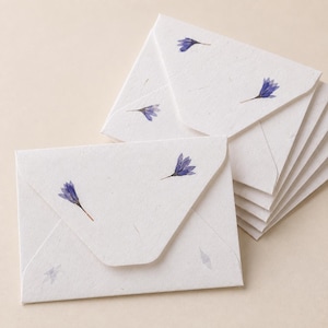 May include: A stack of small, off-white envelopes made from textured paper with visible fibers. Each envelope features a triangular flap and is decorated with small, pressed blue flowers scattered across the surface in a delicate, natural pattern.