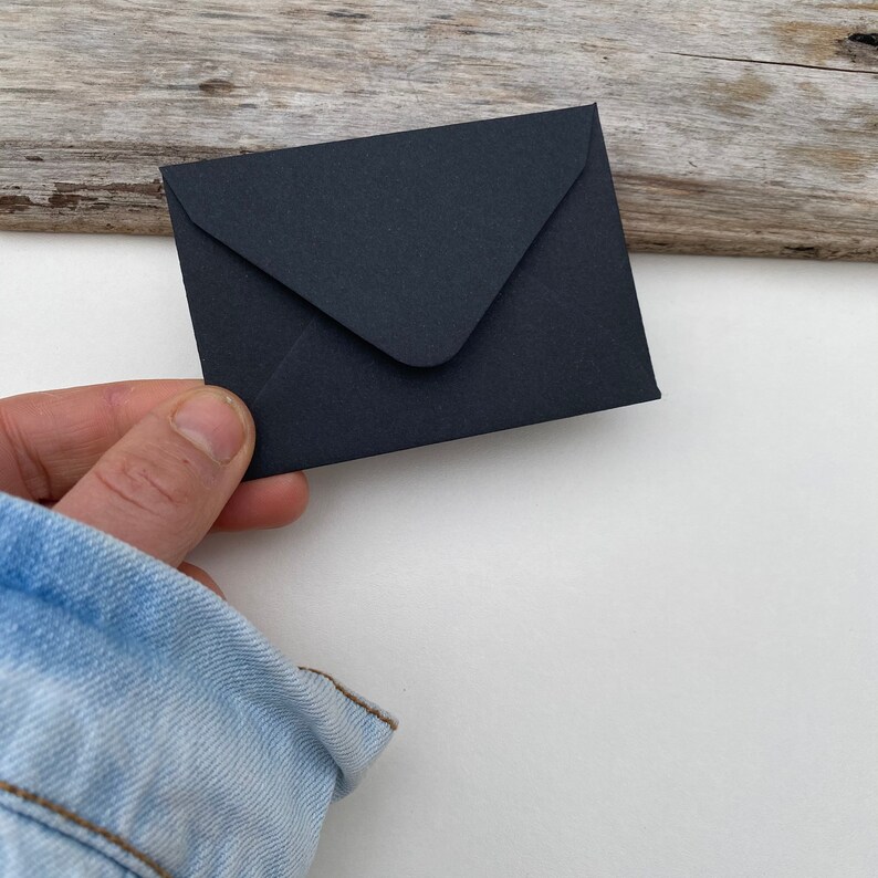May include: A small, black envelope held by a hand. The envelope is rectangular with a triangular flap. The background is a mix of white and weathered wood.