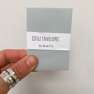May include: A small, pale grey rectangular box with a white band. The band displays the text "Little Envelopes" above "Scraffs" in a simple font. The box is held by a hand, with a silver ring visible on the finger.