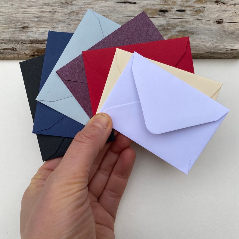 May include: A hand holds a fan of small, colorful envelopes. The envelopes are in various colors, including black, navy, light blue, purple, red, cream, and white. The envelopes are arranged in a semi-circle, showcasing their different hues.