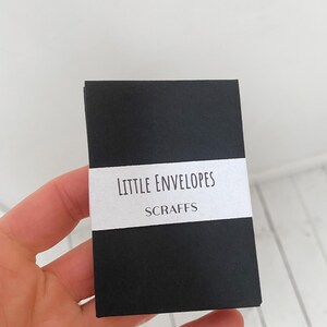 May include: A small, black rectangular package with a white label that reads "LITTLE ENVELOPES" and "SCRAFFS" in a simple font. The package is held in a hand against a white background.