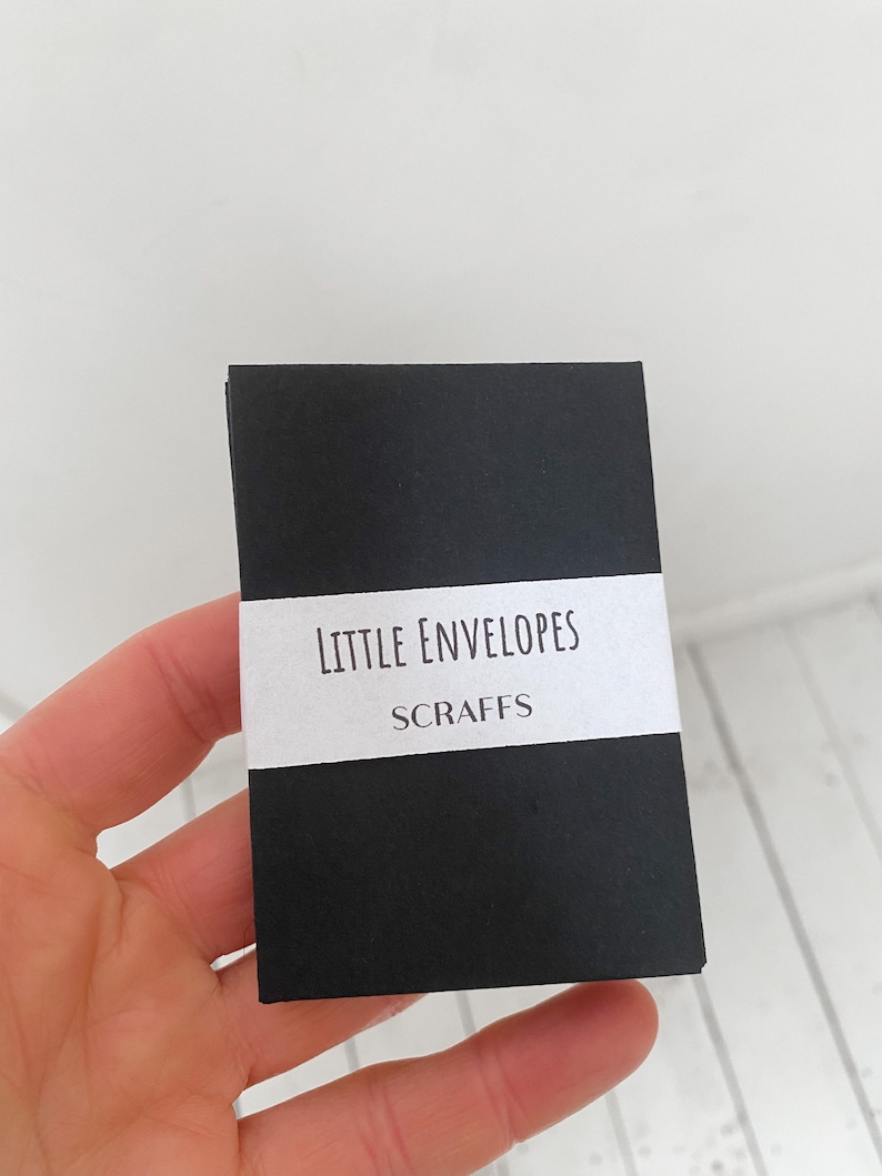 May include: A small, black rectangular package with a white label that reads "LITTLE ENVELOPES" and "SCRAFFS" in a simple font. The package is held in a hand against a white background.