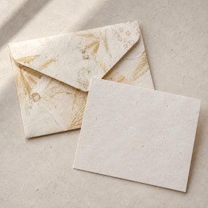 May include: A rectangular off-white card made of textured paper sits next to a matching envelope. The envelope features a pointed flap and is decorated with a subtle, repeating gold botanical print of leaves and flowers across its surface.