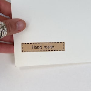 May include: A close-up of a white card with a small, rectangular brown label that reads "Hand made" in a simple, sans-serif font. The label has a dashed border, and the card is held by a hand.