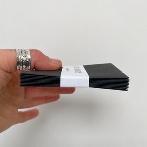 May include: A stack of black notecards held by a hand, with a white band wrapped around the middle. The band features the text "Wild Letter" and "Notes" in a minimalist font. The cards are stacked neatly.