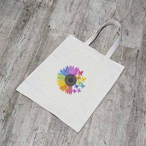 Peut inclure: Sac fourre-tout blanc avec un tournesol arc-en-ciel et des papillons colorés imprimés sur le devant.