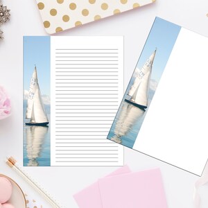 Könnte beinhalten: Ein weißes Segelboot mit einem blauen Streifen am Segel spiegelt sich im Wasser. Das Segelboot steht vor einem blauen Himmel mit weißen Wolken. Das Bild ist ein bedruckbares Briefpapier mit liniertem Papier.