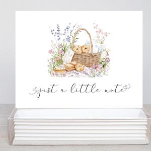 May include: A stack of white note cards featuring a watercolour illustration of a picnic basket filled with pastries and surrounded by flowers. The text "just a little note" is written in cursive below the illustration.