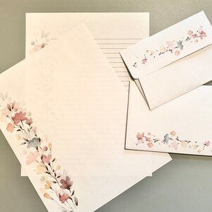 May include: Stationery set featuring white paper and envelopes adorned with a watercolor floral design in soft pink, peach, and blue hues. The set includes lined and unlined sheets, and envelopes.