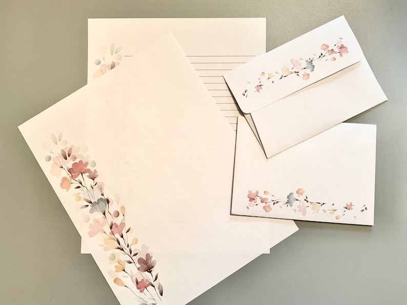 May include: Stationery set featuring white paper and envelopes adorned with a watercolor floral design in soft pink, peach, and blue hues. The set includes lined and unlined sheets, and envelopes.