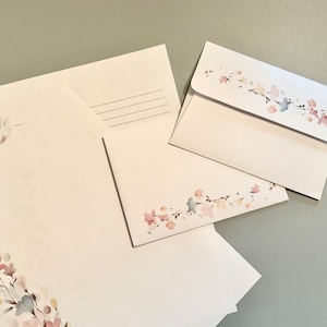 May include: White stationery set featuring floral watercolor designs in pink, blue, and yellow. The set includes envelopes and letter sheets. The envelopes have a floral design on the flap. The stationery is arranged on a gray surface.