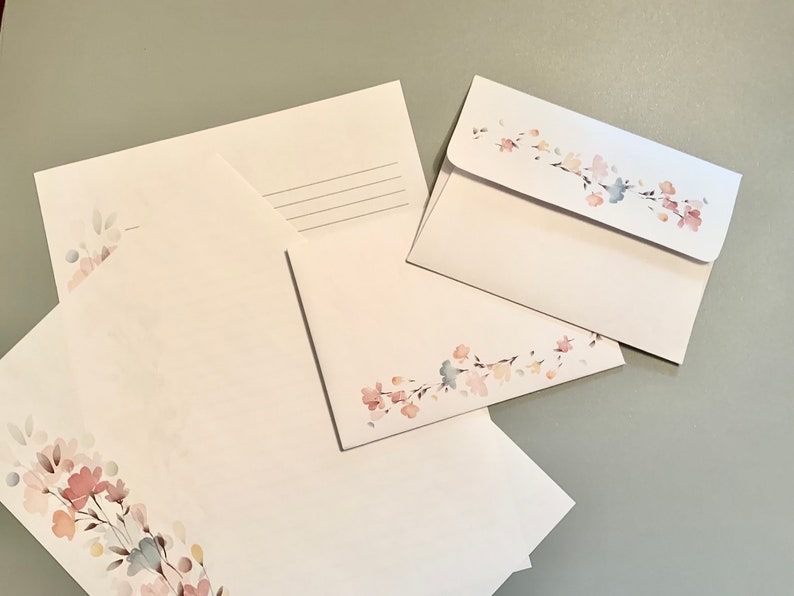 May include: White stationery set featuring floral watercolor designs in pink, blue, and yellow. The set includes envelopes and letter sheets. The envelopes have a floral design on the flap. The stationery is arranged on a gray surface.