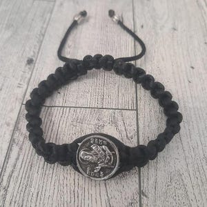Op de afbeelding: Zwart gevlochten armband met een zilverkleurig medaillon met een gegraveerde religieuze figuur. De armband heeft een verstelbare zwarte koordsluiting met zilverkleurige kralen. De armband wordt gepresenteerd op een licht houten oppervlak.