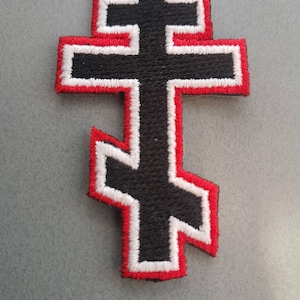 May include: An embroidered patch in the shape of an Eastern Orthodox cross. The cross features a black center with a thick white border and a thin red outer edge. The design includes a top horizontal bar, a longer middle bar, and a slanted bottom bar.