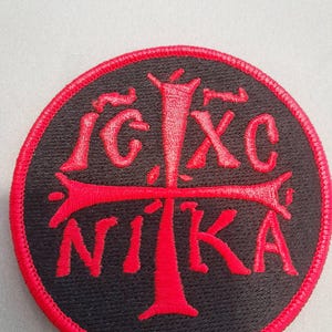 Op de afbeelding: Een ronde geborduurde patch met een zwarte achtergrond, een rode rand en rood stiksel. Het ontwerp toont een centraal kruis met de in rood geborduurde tekst IC XC NIKA rondom het kruis, met IC XC erboven en NIKA eronder.