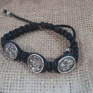 May include: Black braided bracelet featuring three silver-toned circular charms with religious iconography. The bracelet is adjustable with a sliding knot closure. The background is a textured, neutral-toned fabric.