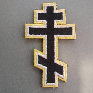 May include: An embroidered patch in the shape of an Eastern Orthodox cross. The cross features a black center with a white border and a yellow outer edge. The design includes three horizontal bars, with the bottom bar positioned at an angle.