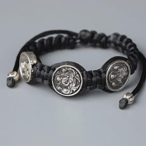 May include: A black braided bracelet with three silver-colored circular charms. The charms feature detailed relief designs. The bracelet has adjustable black cords with silver-colored beads at the ends. The background is a solid gray.