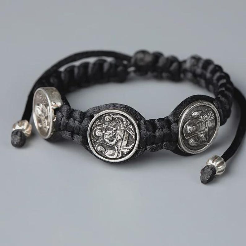 May include: A black braided bracelet with three silver-colored circular charms. The charms feature detailed relief designs. The bracelet has adjustable black cords with silver-colored beads at the ends. The background is a solid gray.