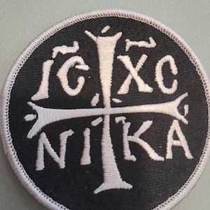 May include: A circular embroidered patch featuring a black background with white stitching. The design displays a central cross and the Greek letters IC XC NI KA arranged around the cross, all enclosed by a white stitched border edge.
