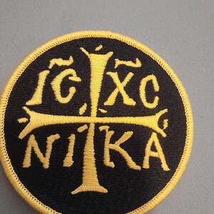 May include: A circular embroidered patch featuring a black background with a yellow border. The design includes a central yellow cross and the yellow Greek letters IC XC NIKA arranged around the cross, representing a religious symbol.