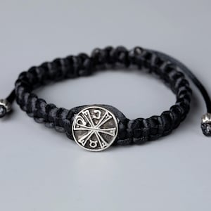 Könnte beinhalten: Schwarz geflochtenes Armband mit silberfarbenem Mittelstück. Das Armband hat ein gewebtes Design und verstellbare Enden. Das Mittelstück hat eine kreisförmige Form mit einem kreuzartigen Symbol.