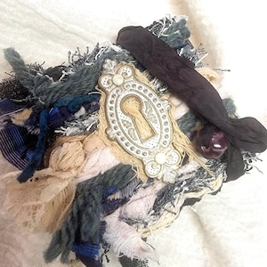 May include: A mixed-media textile art piece featuring a keyhole-shaped embellishment. The artwork incorporates various fabrics in shades of blue, cream, and black, with a dark purple ribbon and a small, round, dark purple bead. The keyhole is framed in a decorative, ornate design.