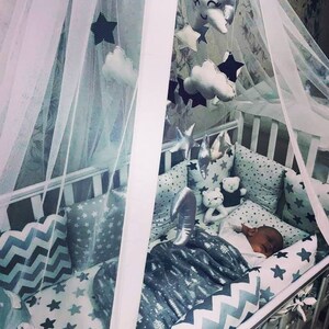 May include: A white crib with a white canopy and a gray and white bedding set. The crib has a mobile hanging above it with clouds and stars. A baby is sleeping in the crib.