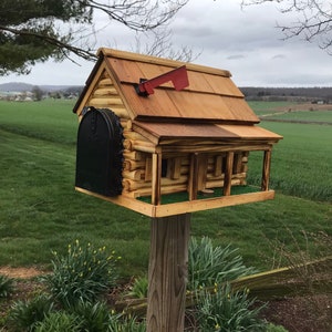 May include: A wooden log cabin style mailbox with a black metal door and a red flag. The mailbox is mounted on a wooden post and is surrounded by green grass.
