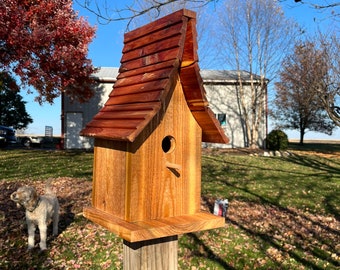 May include: A wooden birdhouse with a red cedar roof, mounted on a wooden post. The birdhouse has a small opening for birds to enter.
