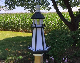 May include: A black and white lighthouse-shaped birdhouse with a white roof and a clear glass window. The birdhouse is mounted on a wooden post and is surrounded by green grass and plants.