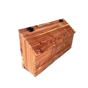 May include: A wooden box with a hinged lid, made of cedar wood with a natural reddish-brown color. The box has black metal hinges.