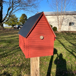 Op de afbeelding: Een rood houten vogelhuisje met een zwart dak, een ronde opening en een houten paal. Het vogelhuisje staat op een grasveld met een schaduw die door de zon wordt geworpen.