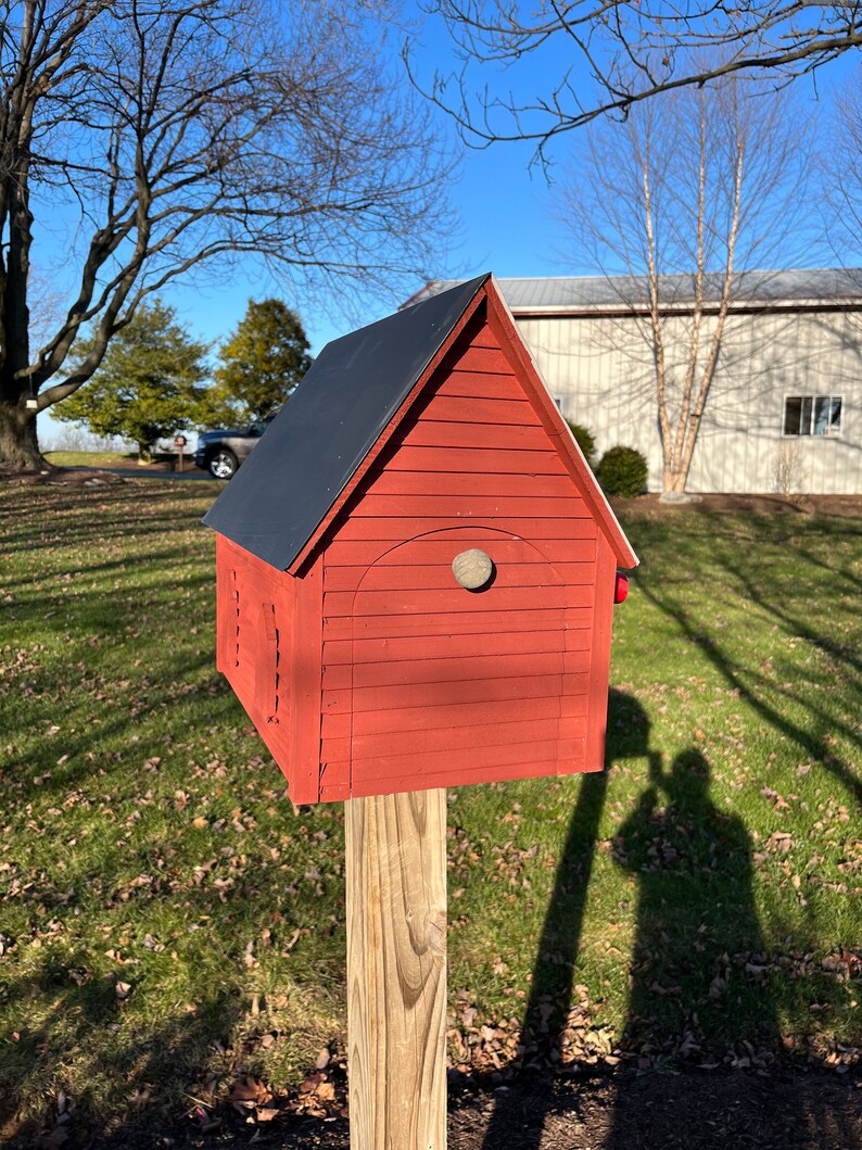 Op de afbeelding: Een rood houten vogelhuisje met een zwart dak, een ronde opening en een houten paal. Het vogelhuisje staat op een grasveld met een schaduw die door de zon wordt geworpen.