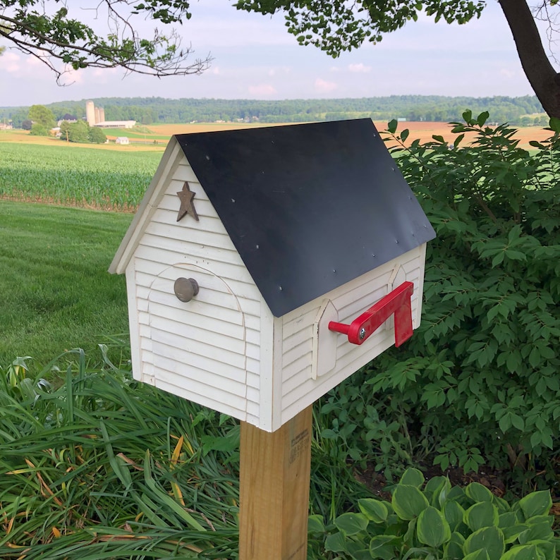 Op de afbeelding: Witte houten brievenbus in de vorm van een huis met een zwart dak en een rode vlag. De brievenbus heeft een ster aan de voorkant en een kleine deur.