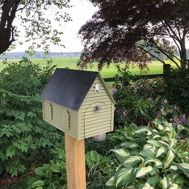 Op de afbeelding: Een kleine, groene, houten huisvormige brievenbus met een zwart dak en een witte ster aan de voorkant. De brievenbus is gemonteerd op een houten paal en is omringd door groene planten.